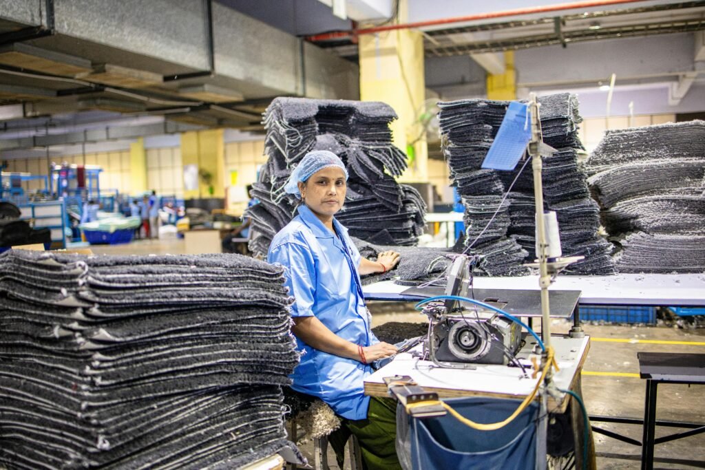 A factory worker operates a sewing machine surrounded by textile stacks in an industrial setting.